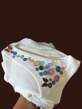 Load image into Gallery viewer, A folded white ribbed knit ladies tank tops, likely a baby bodysuit or shirt, decorated with colorful buttons and butterfly appliqués, held against a dark blue background.

